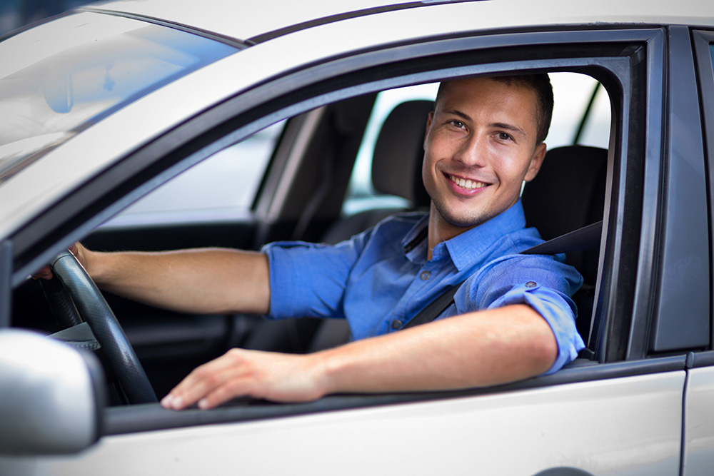 A man smiling and sitting in the driver's seat of a white car, with his arm resting on the window sill.