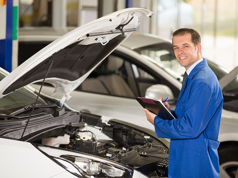 A mechanic in a blue jumpsuit smiles while inspecting a vehicle's engine, holding a clipboard with a pen in hand.