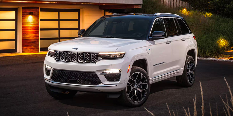 White Jeep Grand Cherokee SUV parked in a modern residential driveway at dusk, with exterior lights on and a contemporary garage in the background.