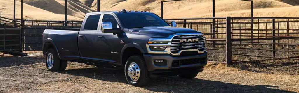 Dark gray 2026 Ram 3500 heavy-duty pickup truck parked on a dirt path in a rural setting, with fencing and rolling hills in the background.