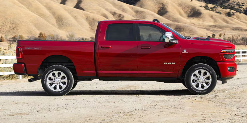 Side view of a red Ram pickup truck parked on a dirt road with hills and a white fence in the background