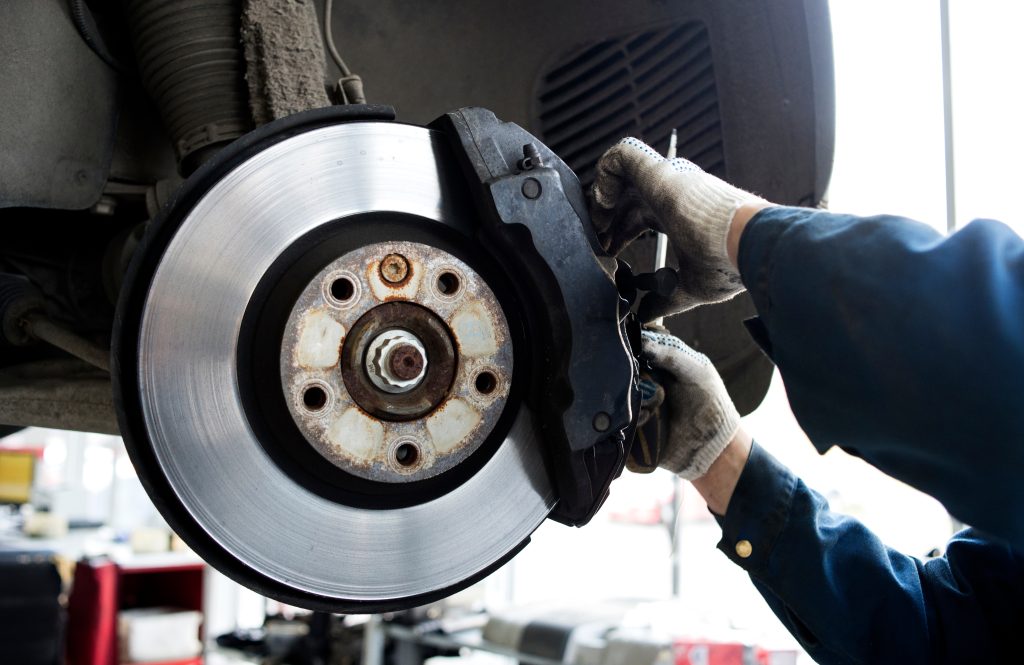 Close-up view of a car brake rotor and caliper being inspected or repaired by a mechanic wearing gloves in an automotive workshop.