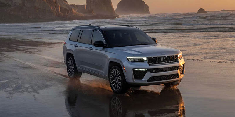 Silver SUV driving along a wet beach shoreline at sunset, with ocean waves, rocky cliffs, and warm golden lighting reflecting on the sand.