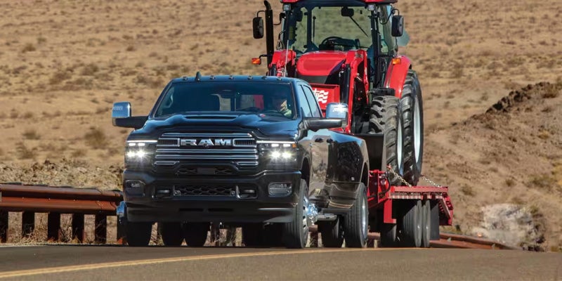 Black RAM pickup truck towing a large red tractor on a trailer through a rural landscape.